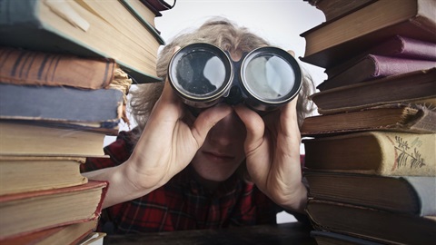 Person with binoculars peering past piles of books